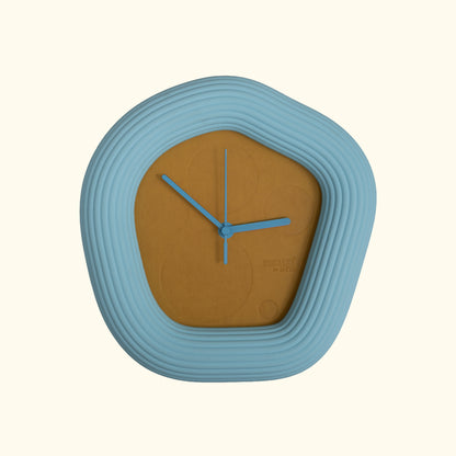 A modern Wallie wall clock with a light blue concentric rounded square frame and a warm brown center, shown against a solid cream background. Made by Ura Watches / EON TIME