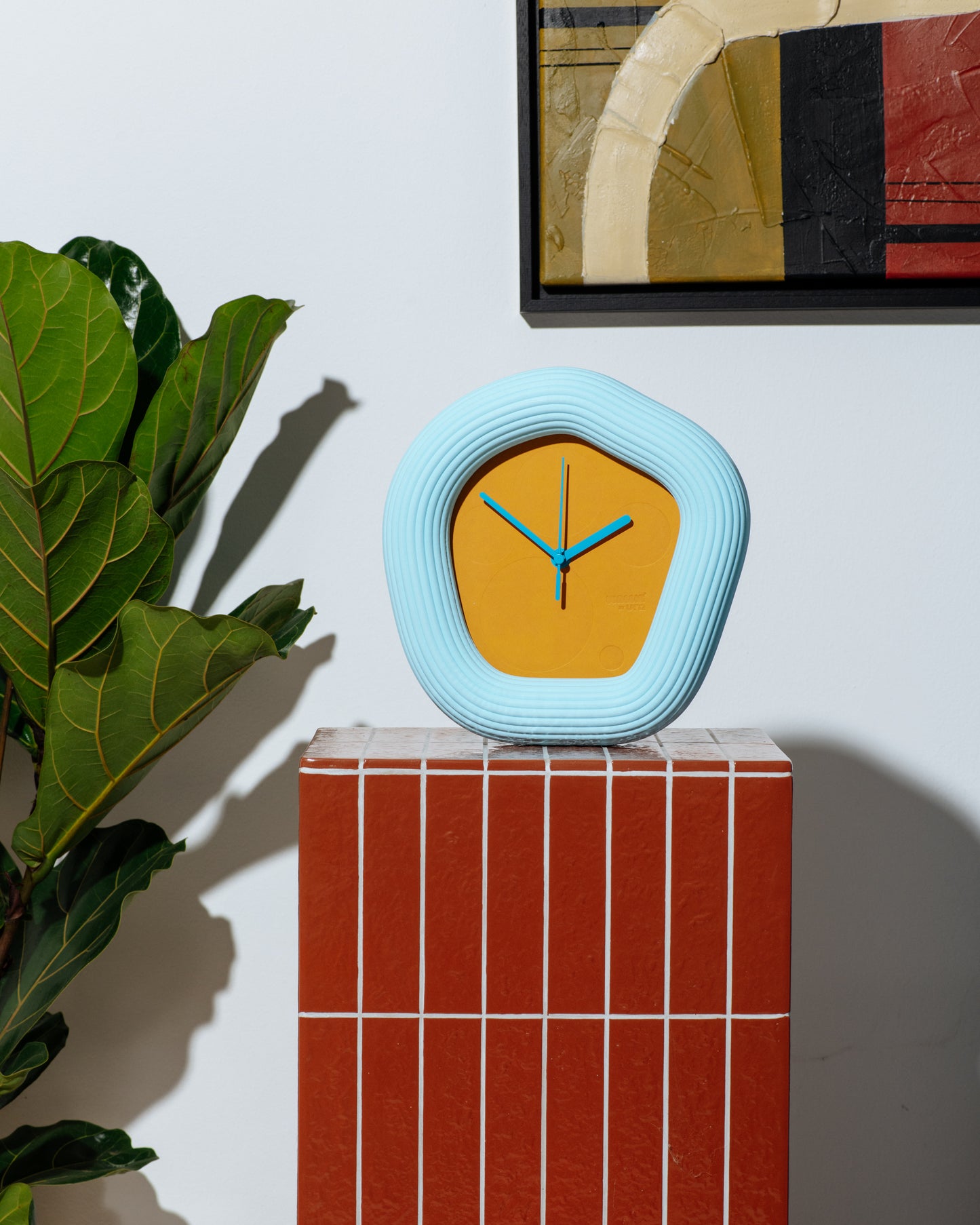 A modern, organic-shaped wall clock with a light blue ribbed frame and a mustard yellow face, displayed on a red tiled pedestal next to a green plant. Made by Ura Watches / EON TIME
