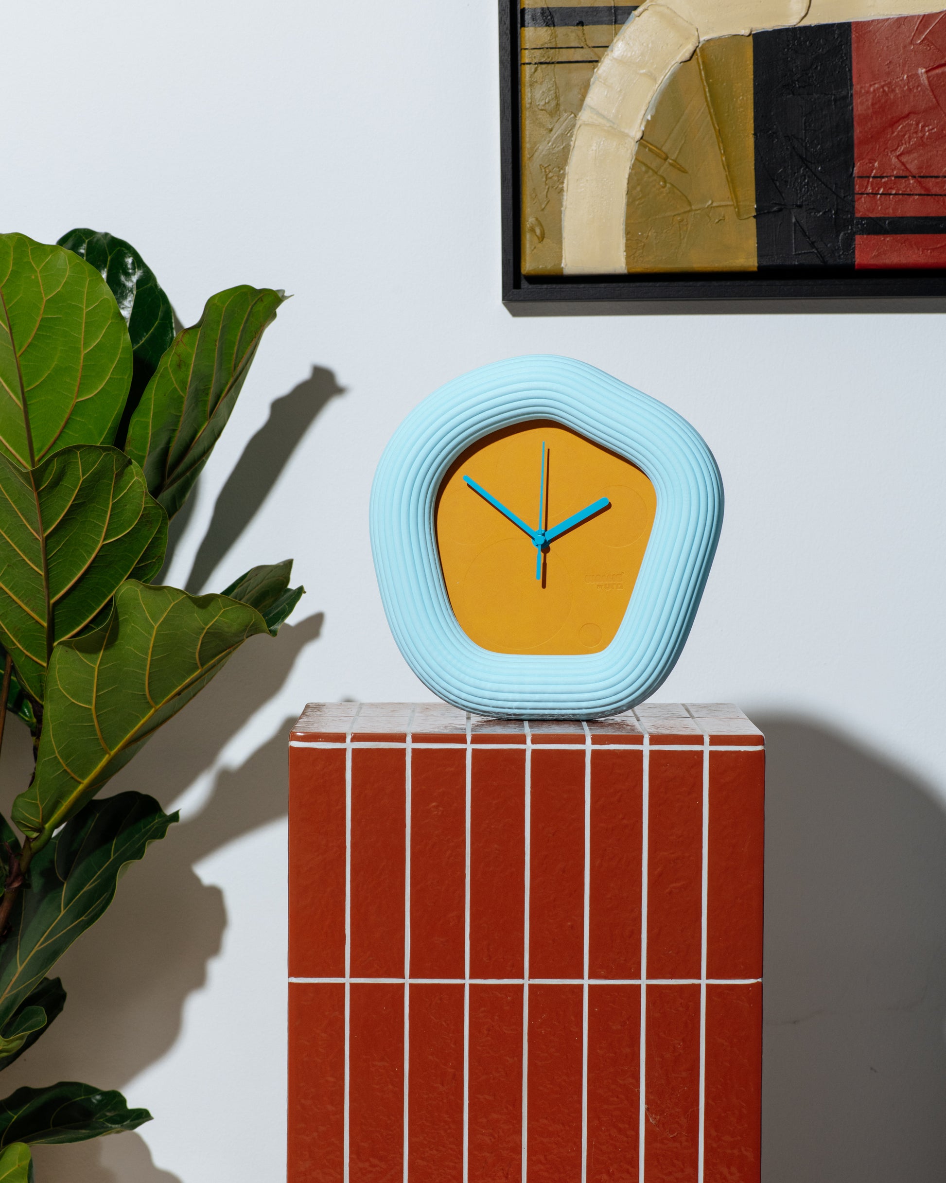 A modern, organic-shaped wall clock with a light blue ribbed frame and a mustard yellow face, displayed on a red tiled pedestal next to a green plant. Made by Ura Watches / EON TIME
