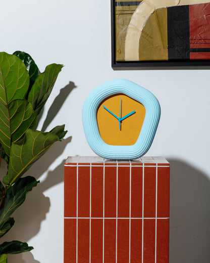 A modern, organic-shaped wall clock with a light blue ribbed frame and a mustard yellow face, displayed on a red tiled pedestal next to a green plant. Made by Ura Watches / EON TIME