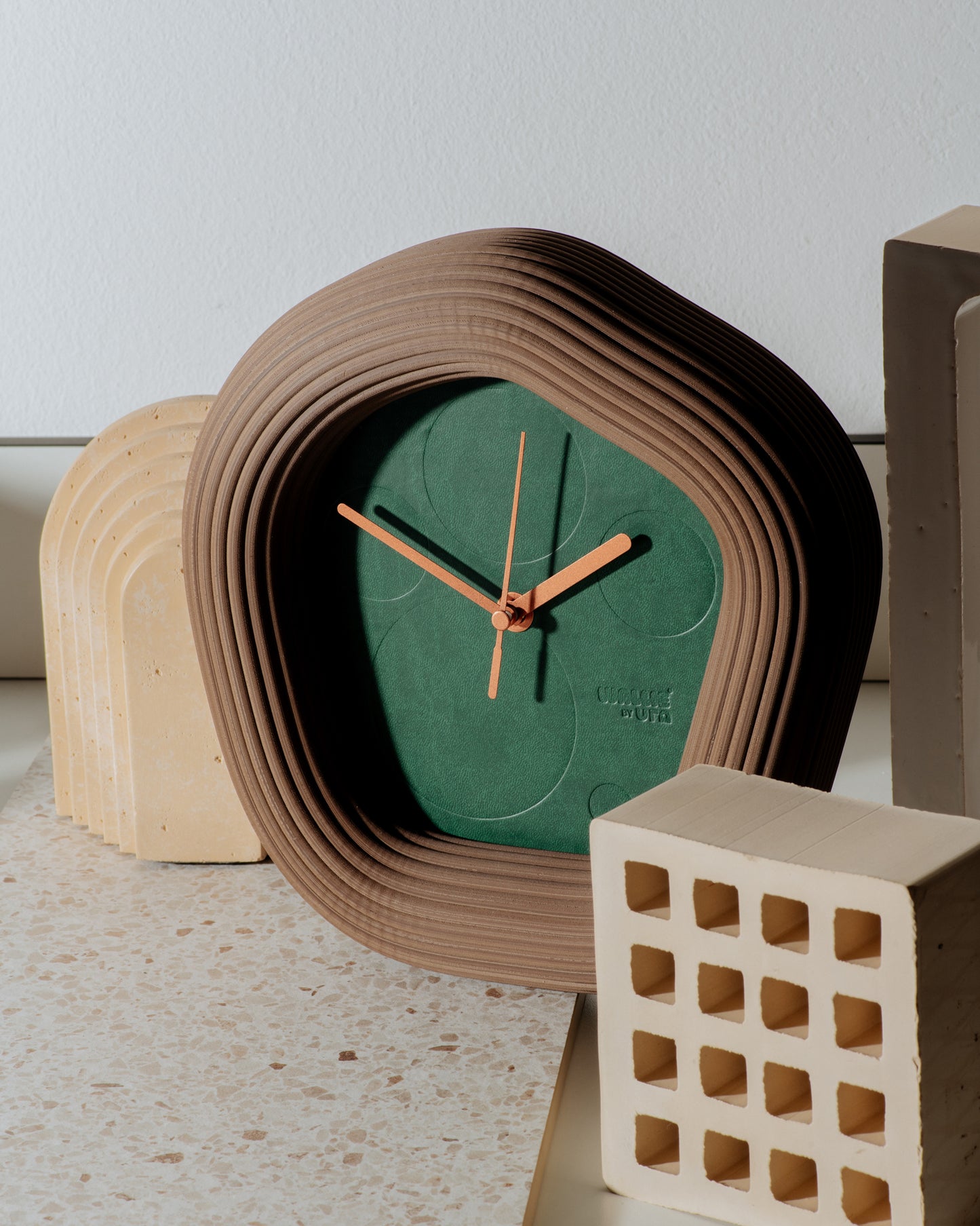A sculptural Wallie wall clock with a terracotta-brown wavy frame and a dark green face, styled on a light stone surface with neutral decorative blocks. Made by Ura Watches / EON TIME