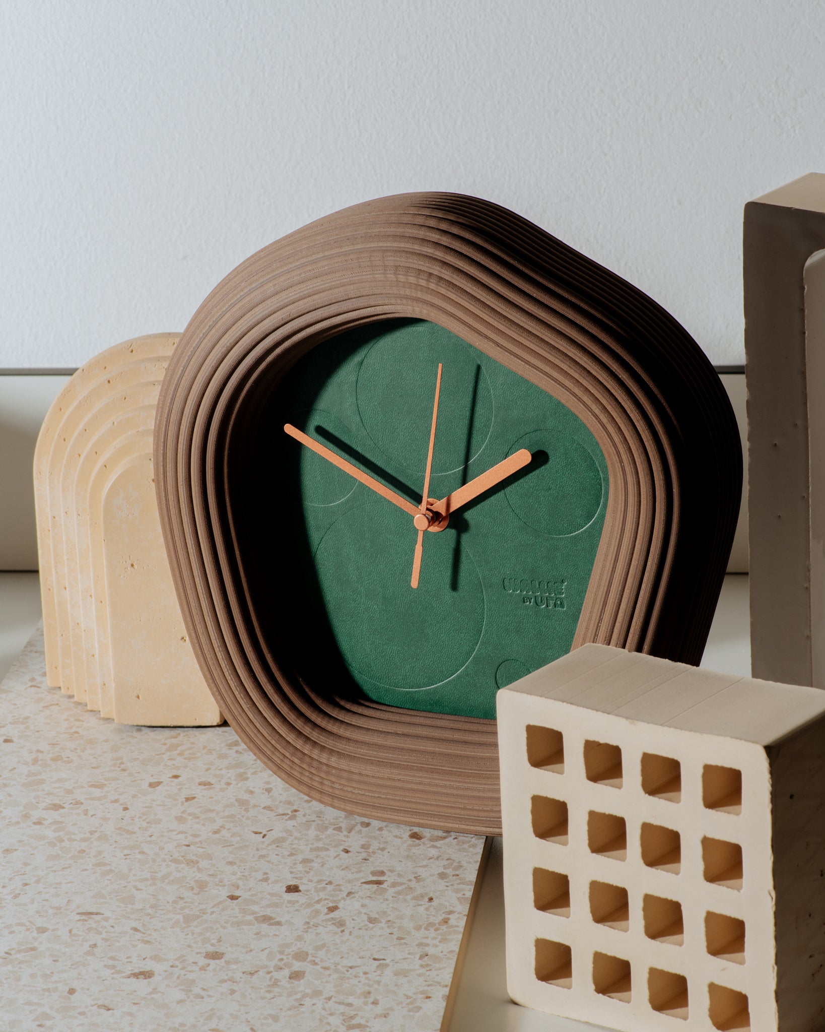 A sculptural Wallie wall clock with a terracotta-brown wavy frame and a dark green face, styled on a light stone surface with neutral decorative blocks. Made by Ura Watches / EON TIME