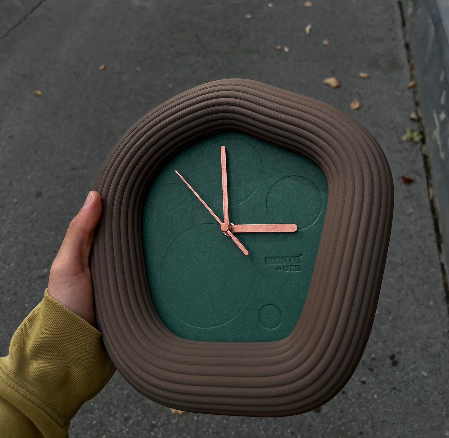 Person holding a unique Wallie clock with a textured brown casing against a concrete background. Made By Ura Watches / EON TIME