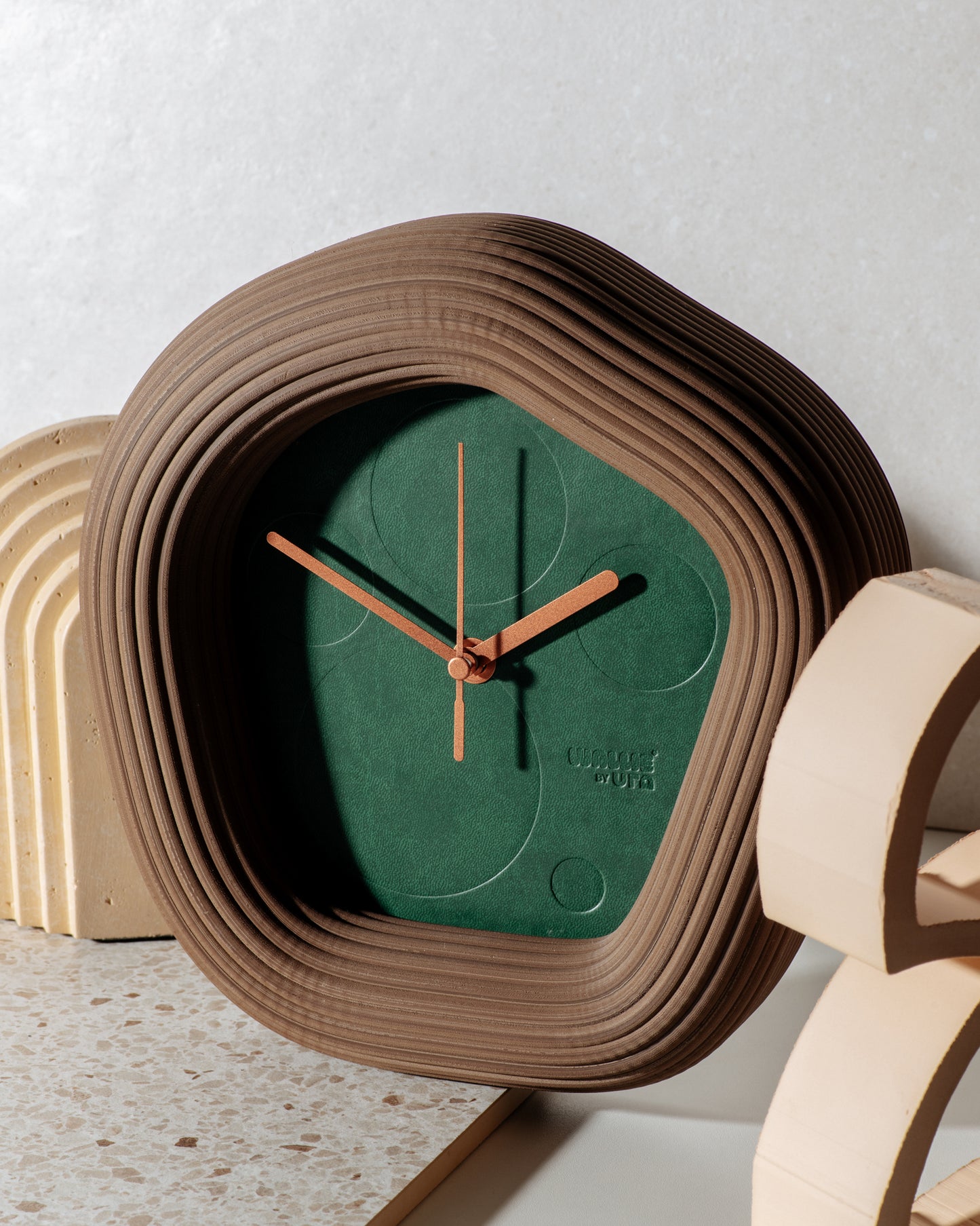 A modern round Wallie wall clock with a sculptural layered brown frame and a dark green dial, styled on a terrazzo surface with light wood arch props against a textured light gray wall. Made by Ura Watches / EON TIME