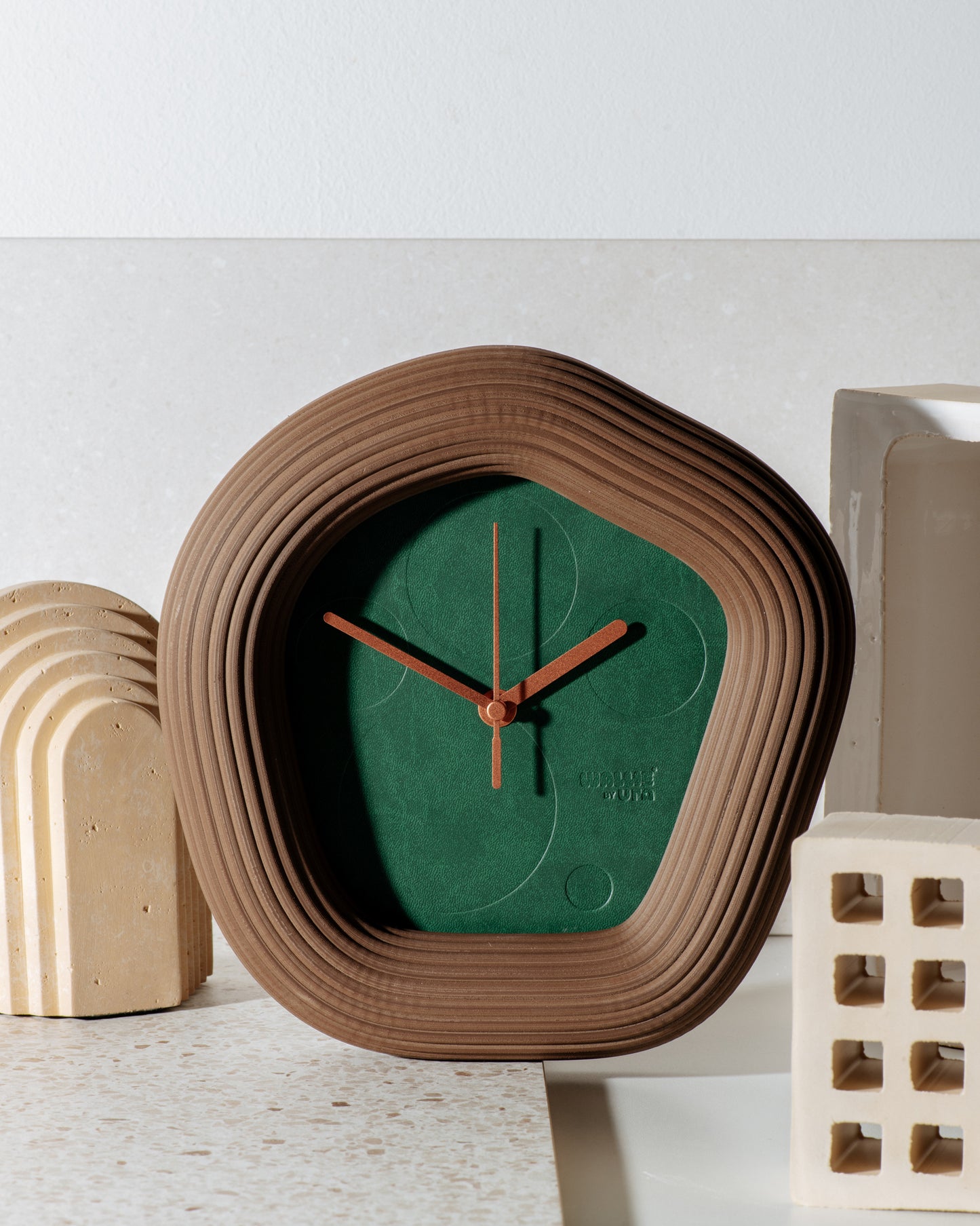 A modern organic-shaped Wallie wall clock with a textured brown concentric frame and a green dial, styled on a light stone surface with neutral geometric props. Made by UraWatches / EON TIME