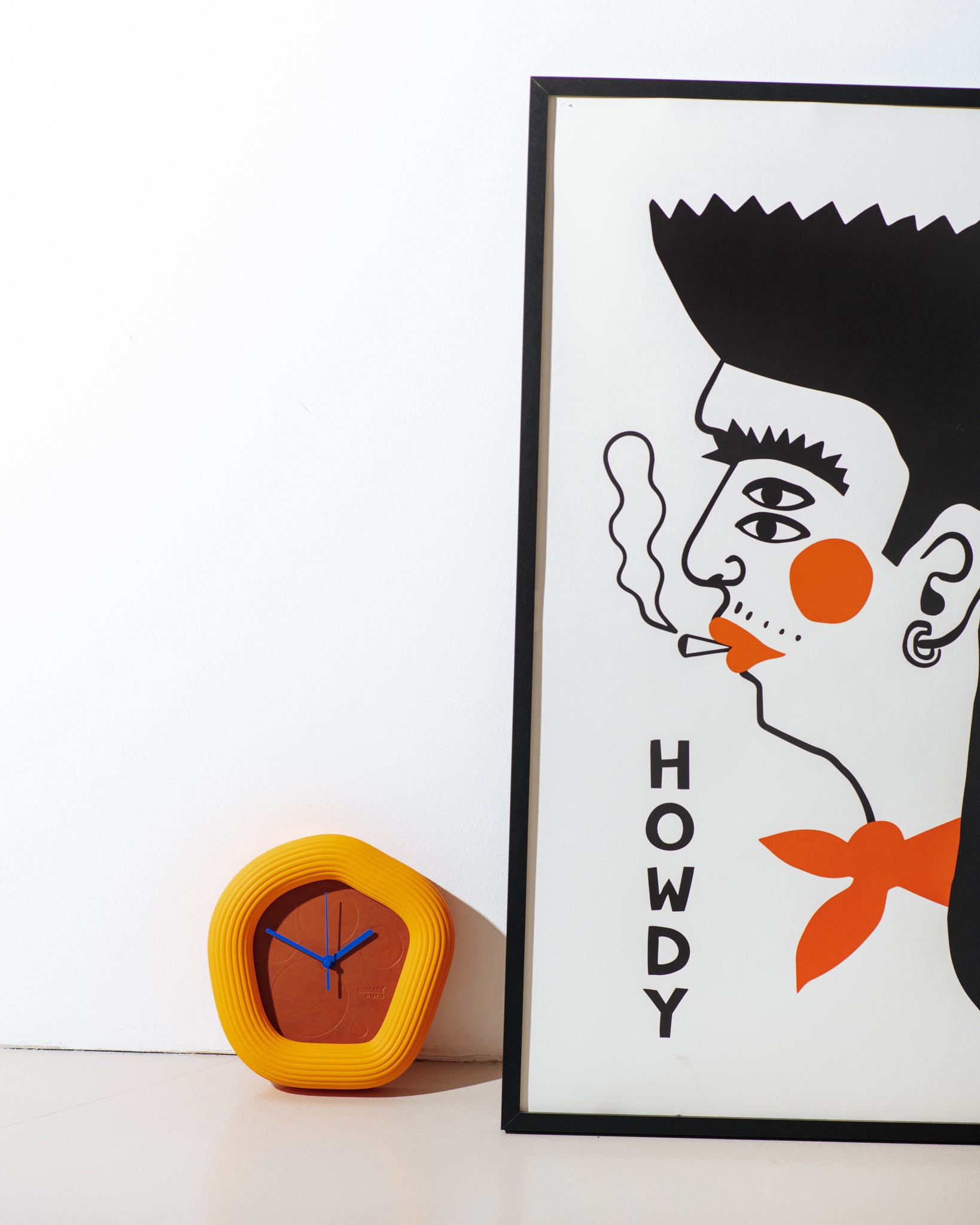 A framed art print featuring a stylized black and orange illustration of a man's face with the word 'HOWDY' printed vertically, displayed next to a yellow modern wall clock on a white surface against a white wall. with orange Wallie by it. made by Ura Watches / EON TIME