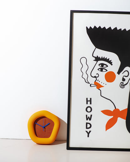 A framed art print featuring a stylized black and orange illustration of a man's face with the word 'HOWDY' printed vertically, displayed next to a yellow modern wall clock on a white surface against a white wall. with orange Wallie by it. made by Ura Watches / EON TIME