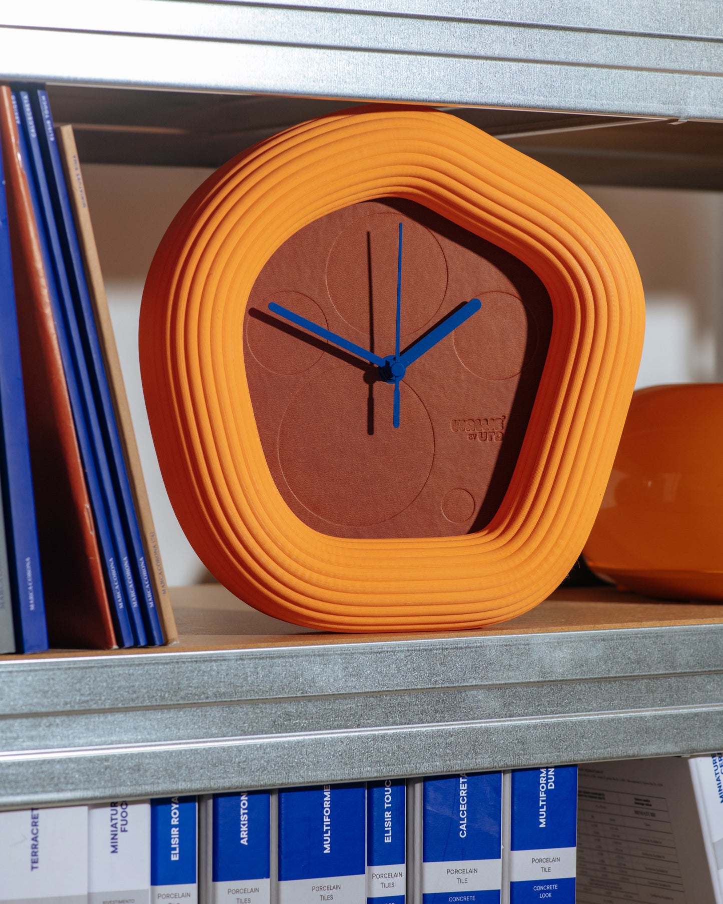 Orange Wallie clock with blue hands on a shelf with books