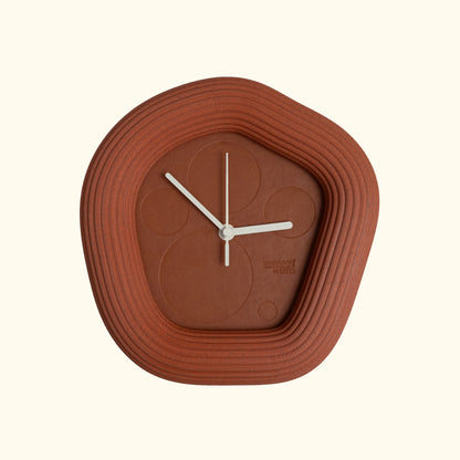 An organic-shaped terracotta Wallie wall clock with concentric ridged outer rings and minimalist white hands, shown against a solid cream background.