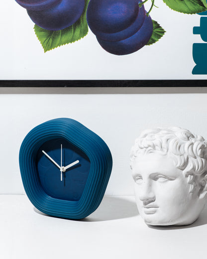 A modern blue concentric-ripple Wallie clock with a dark blue face and silver hands, styled on a white surface next to a white classical bust sculpture and a framed print of plums. Made by Ura Watches in Croatia