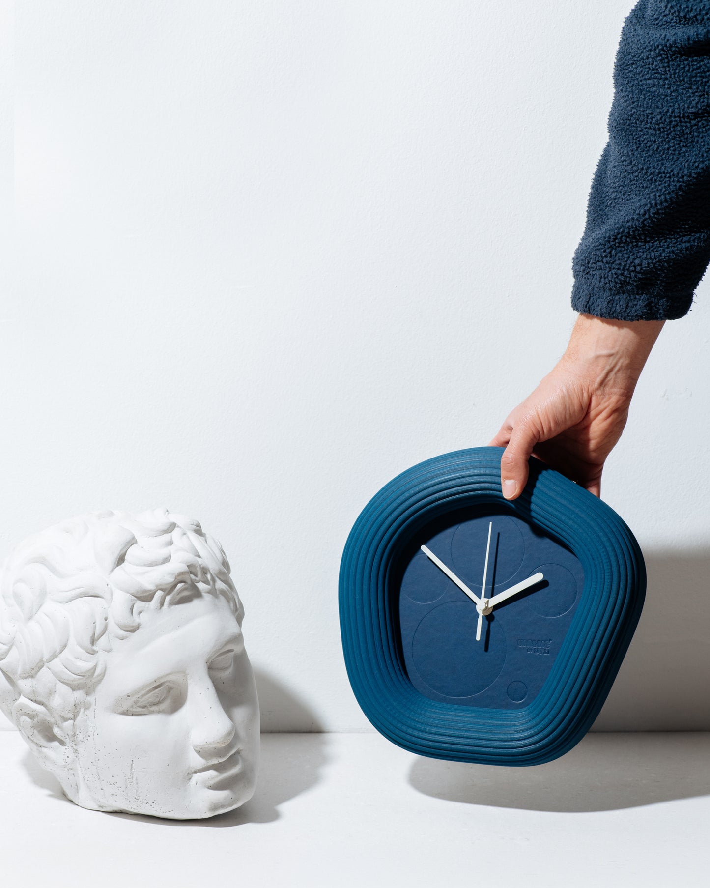 A hand in a dark blue fleece sleeve holds a dark blue, uniquely shaped Wallie clock next to a white classical bust sculpture on a clean white background. Made by Ura Watches from Croatia