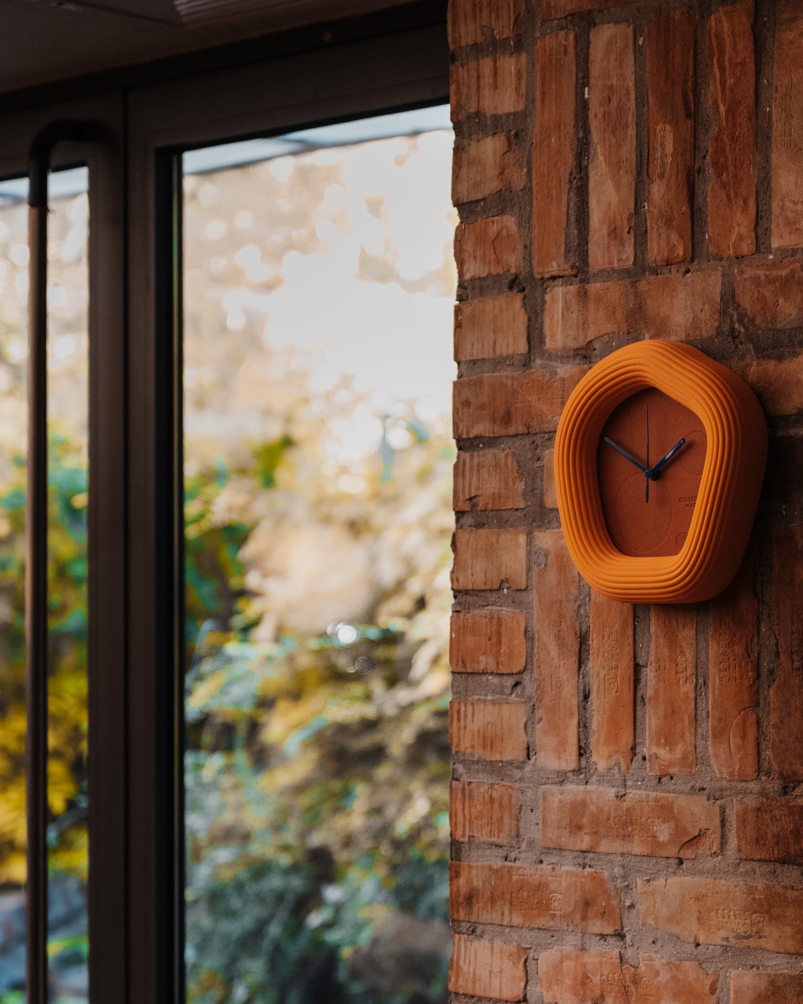 Orange Wallie wall clock on a brick wall with a blurred outdoor scene in the background. Made by Ura Watches / EON TIME